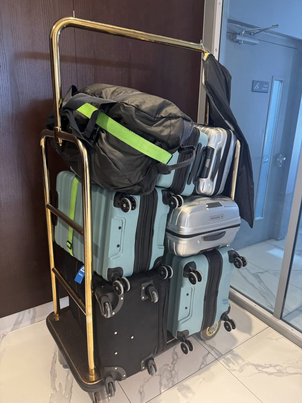 A luggage trolley in an apartment lobby, stacked with four wheeled suitcases and a duffel bag.